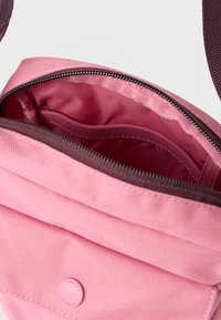 Ροζ υφασμάτινη τσάντα με κλείσιμο με φερμουάρ. Το εσωτερικό διαθέτει ομαλή επένδυση και ξεχωριστή θήκη, συνοδευόμενα από μπορντό ταινία και λεπτομέρεια λογότυπου.