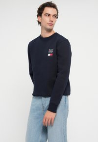 Námornícky sveter s rebrovaným výstrihom, s bielym a červeným vyšitým logom na hrudi. Spárovaný s svetlo modrými denimovými džínsmi.