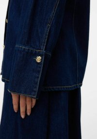Main reposant sur le côté, portant une veste en denim bleu marine avec un bouton de manchette texturé doré visible et une jupe assortie.