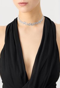 Clear crystal choker with an assortment of round and oval stones, set in silver-toned metal, worn with a black draped fabric top.