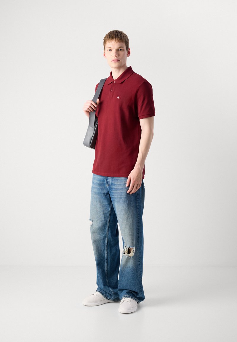 Młody mężczyzna w czerwonej koszulce polo i podartych niebieskich dżinsach, trzymający szary torbę na ramieniu, stojący na tle jasnego, gładkiego tła.