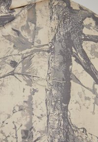 Maastokuvioitu kangas, jossa puun ja oksien kuviot harmaan sävyissä beigen pohjalla, sekä hienovarainen kirjailtu logoyksityiskohta.