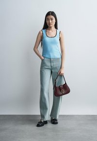 Fiatal nő világoskék ujjatlan felsőt, világos farmernadrágot és fekete loafereket visel, kezében sötétvörös kézitáska, szürke egyszínű háttér előtt.