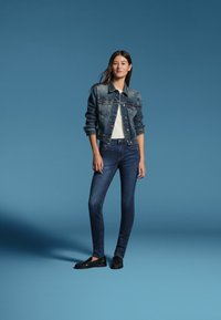 Frau steht vor blauem Hintergrund, trägt eine Jeansjacke, ein weißes Top, dunkle Jeans und schwarze Loafers.