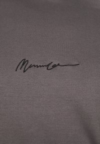 Tummanharmaa kangas, jossa mustalla käsinkirjoitetulla tekstillä on brodeerattu sana "Mannica" lähellä keskikohtaa.