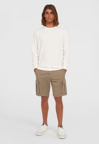 Vaalean beige huppari pitkillä hihoilla, yhdistettynä khaki cargo-shortsien kanssa, joissa on sivutaskut. Yhdistetty valkoisiin lenkkareihin.
