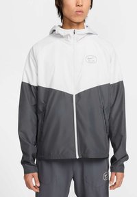 Herre vindjakke i hvid og mørkegrå med lynlåslukning, hætte og sidelommer. Materialet ser glat ud med et sporty design.
