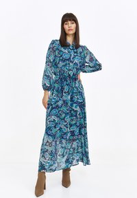 Model przyjmuje pozę w długiej, zwiewnej sukience w wzory paisley w odcieniach niebieskiego i zielonego, zestawionej z brązowymi botkami na obcasie na tle białego tła.