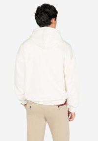 Sudadera blanca con un bolsillo canguro en la parte frontal y una capucha grande, combinada con pantalones de un beige claro, que muestran una tela texturizada y un ajuste relajado.