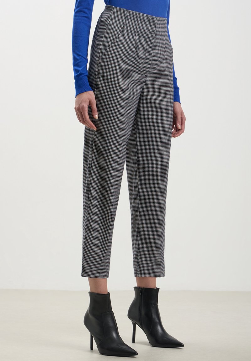 Femme portant un pantalon noir et blanc à carreaux longueur cheville, un haut bleu à manches longues et des bottines noires à talons hauts debout.