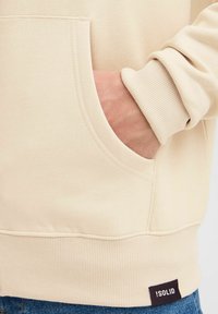 Beige Kapuzenpullover mit Kängurutasche, gerippten Bündchen und Saum. Verfügt über einen strukturierten Stoff und ein kleines Markenlabel am unteren Saum.