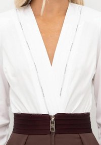 Maglietta bianca a maniche lunghe con scollo a V profondo e trama verticale a costine, abbinata a una fascia plissettata color vinaccia e dettaglio con cerniera argento.