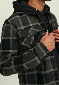 Camicia di flanella a quadri nera e grigia con bottoni, due tasche sul petto e una felpa con cappuccio nera sotto. Tessuto testurizzato.