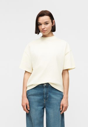 Joven con cabello corto y castaño, vistiendo una blusa de manga corta suelta de color crema y vaqueros de mezclilla azul, sobre un fondo liso.