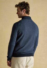 Navy zila adīta džemperis ar augstu apkakli, ribotām piedurknēm un apakšu, ar teksturētu virsmu. Valkāts virs krēmkrāsas kordūru biksēm.