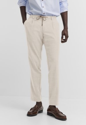 Mann trägt beige Kordhosen, ein blau gestreiftes Hemd mit hochgekrempelten Ärmeln und braune Ledermokassins, steht mit den Händen teilweise in den Taschen.