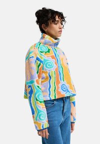 Φλις πουλόβερ σε παστέλ χρώματα με στροβιλιζόμενα και γραμμικά σχέδια, ψηλός γιακάς και κοντό μήκος. Συνδυάζεται με μπλε τζιν.