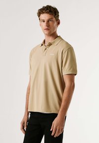 Beige Poloshirt mit kurzen Ärmeln und klassischem Kragen. Verfügt über ein dezentes Logo auf der Brust und eine lockere Passform, kombiniert mit schwarzen Hosen.