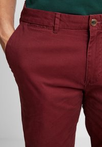 Pantaloni in cotone borgogna con zip, chiusura a bottone e tasche laterali. Il tessuto ha una trama liscia con un leggero lucido.
