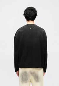 Melna sporta džemperis ar garām piedurknēm, apaļu kaklu un divām metāla acīm muguras daļā. Audums ir gluds ar brīvu piegriezumu.