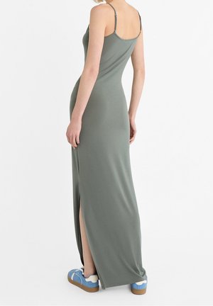 Robe maxi vert olive avec fines bretelles, dotée d'une fente latérale. Associée à des baskets bleues, mettant en valeur des textures et couleurs contrastées.