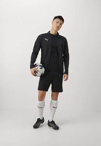 Zwarte sportjas met ritssluiting en logo, zwart T-shirt, korte broek, witte sokken met logo en zwarte voetbalschoenen; houdt een voetbal vast.