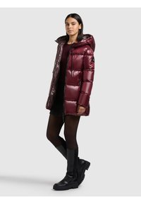 Burgundiškos spalvos pūkinė striukė su kapišonu, blizgiu paviršiumi, užtrauktuku ir dviem šoninėmis kišenėmis. Juodi iki kelių siekiantys batai užbaigia įvaizdį.