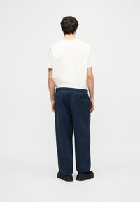 Άτομο που φοράει λευκή κοντομάνικη μπλούζα και φαρδιά σκούρα μπλε παντελόνια με πίσω τσέπη, στέκεται γυρισμένο πλάτη σε απλό φωτεινό φόντο.