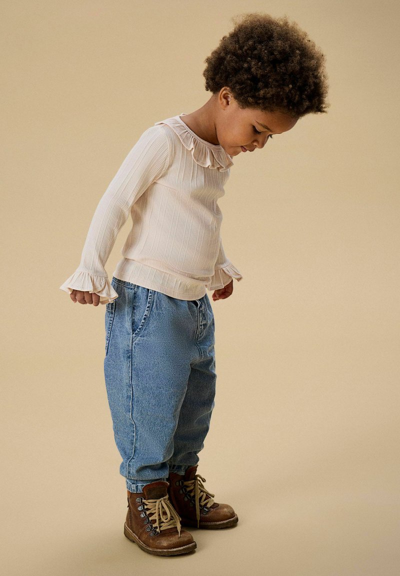 Lys kremfarget langermet topp med rysjekrage, kombinert med løstsittende blå jeans og brune snørebåndsstøvler. Enkelt og teksturert design.