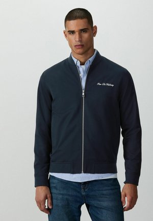 Mann trägt eine marineblaue Jacke mit Reißverschluss über einem blau-weiß gestreiften Hemd und Jeans, steht vor einem schlichten hellen Hintergrund.