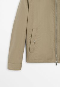 Manica di giacca beige e lato inferiore con cerniera verticale sul davanti e tasca obliqua con bottone a pressione su uno sfondo uniforme.