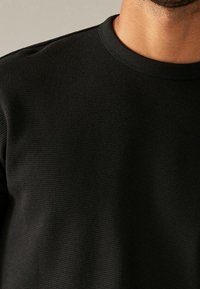 Chemise noire texturée à manches longues, avec un col rond et un motif côtelé subtil, présentée de la zone de la poitrine.