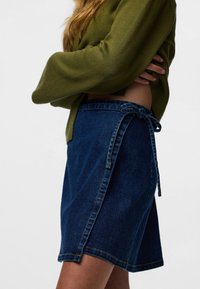 Tumesinise tooniga teksaseelik, millel on küljeäärsed sõlmitavad detailid, sirge lõige ja sõlmevahemikuga ääris. Tekstuuriga materjal on nähtav.