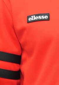 Punane polo särk, millel on musta tikandiga "ellesse" logo ja kolm musta triipu varruka peal. Valmistatud tekstureeritud kangast.