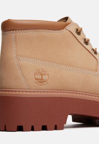 Stivale in pelle impermeabile color beige con colletto alla caviglia marrone, suola in gomma rossa a contrasto e dettagli di cucitura visibili. Logo impresso di lato.