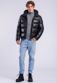 Jonge man die een zwarte pufferjas, een grijze sweater met rits, lichtblauwe spijkerbroek en zwarte veterschoenen draagt, staand tegen een effen achtergrond.