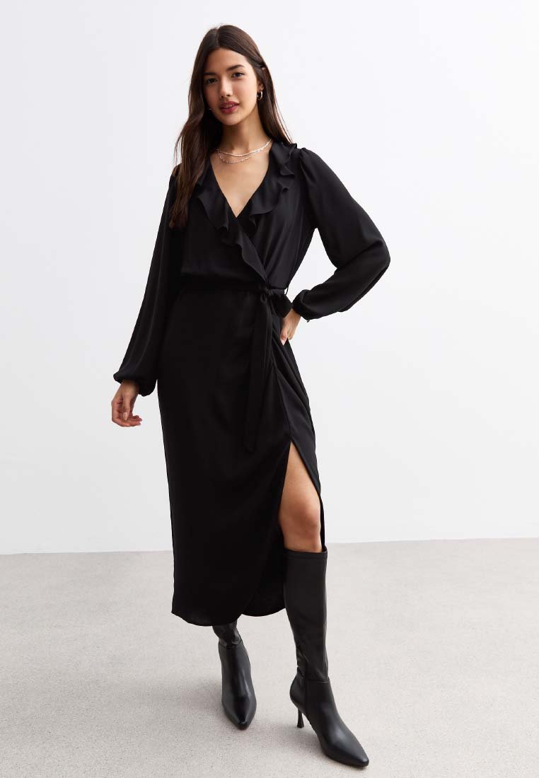 Robe portefeuille noire à manches longues avec volants, fente sur le côté et ceinture à la taille, fabriquée en matière lisse. Portée avec des bottes noires jusqu'aux genoux.