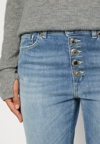 Personne portant un jean bleu clair avec quatre boutons décoratifs en argent et un pull gris côtelé avec des trous pour les pouces.