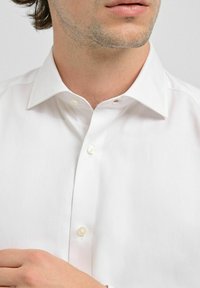 Vit skjorta med klassisk krage, som har pärlknappar och en mjuk textur. Närbild på övre delen, som framhäver knäppt placket.