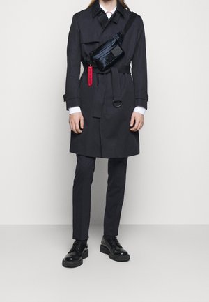 Czarny trench coat z paskiem w talii, zestawiony z wąskimi spodniami i czarnymi sznurowanymi butami. Czarne torby na ramię i czerwony detal z metką.