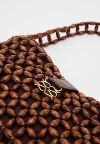 Τσάντα με καφέ ξύλινες χάντρες, χρυσό διακοσμητικό κούμπωμα και καφέ δερμάτινο λουράκι σε λευκό φόντο.