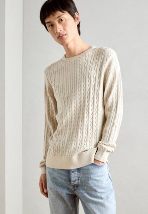 Beige kabelgebreide trui met ronde hals, geribde manchet en zoom. Gecombineerd met lichtblauwe spijkerbroek en weergegeven tegen een neutrale achtergrond.