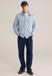 Cămașă albastru deschis, cu mâneci lungi și butoniere, având două buzunare frontale, asociată cu blugi albaștri închis și pantofi maronii, completată cu o șapcă bleumarin.