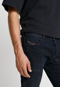 Tenké tmavě modré džíny ze denimu s efektem opotřebení, pěti kapsami a nenápadnou značkou u zadní kapsy.