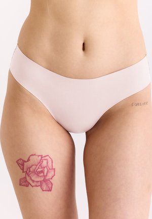 Frau trägt hellrosa Unterwäsche, zeigt Rosen-Tattoo am linken Oberschenkel und "FOREVER"-Tattoo an der rechten Hüfte.