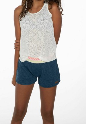 Młoda osoba z kręconymi włosami, ubrana w bezrękawnik z białym wzorem w cętki lamparta oraz niebieskie, casualowe szorty na białym tle.