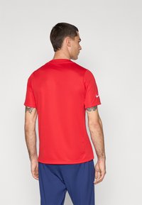 Piros sportpóló rövid ujjakkal, könnyű anyagból készült. Kerek nyakkivágással rendelkezik, és a bal ujján található a Nike logó.
