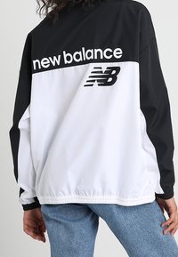 Czarno-biała kurtka wiatrówka z logo "new balance" na plecach, elastycznymi mankietami i luźnym krojem. Teksturowany materiał o sportowym wzornictwie.