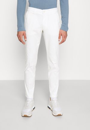 Mand iført hvide slim-fit bukser, hvide sneakers og en blå langærmet skjorte, stående mod en ensartet lys baggrund.