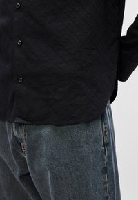 Mustrood mustriga mustreeritav särk, millel on tekstuur, nööbid, kõver hem ja tõstetud logo allosas. Kombineeritud laia sinise teksapükstega.